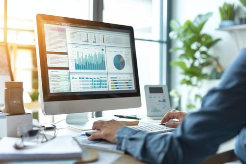 Na obrázku je počítačový monitor zobrazujúci grafy a tabuľky. Počítač sa nachádza na pracovnom stole, kde je aj klávesnica, myš a ďalšie kancelárske potreby. V pozadí sú vidieť rastliny a svetlé okno.