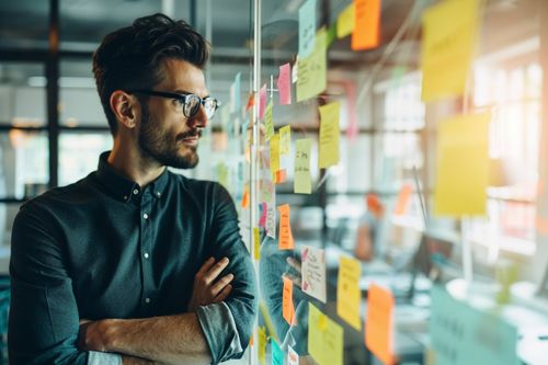 Muž s kríženými rukami, ktorý pozorne hľadí na farebné poznámkové lístky nalepené na sklenenej stene.