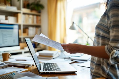 Osoba stojí pri stole s otvoreným laptopom, drží niekoľko papierov. Na stole sú rozložené ďalšie dokumenty a hrnček. V pozadí je polica s knihami a lampa.