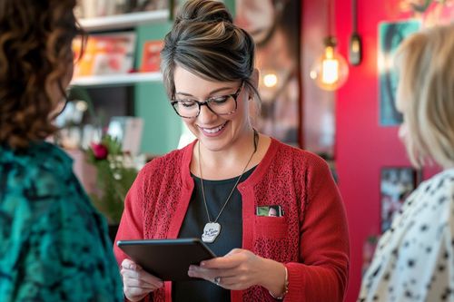 Osoba v červenom svetri s okuliarmi drží tablet a usmieva sa. Na pozadí sú rozmazané farebné steny a dekorácie.