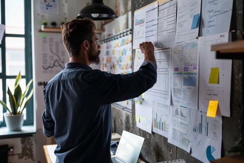 Osoba stojí pri stene pokrytej grafmi a dokumentmi, píše alebo upravuje niečo na papieri. V pozadí je vidieť počítač a kvetinu na parapete.